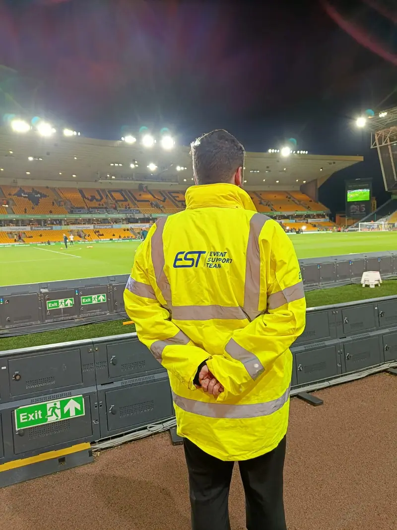 An EST steward in a hi-vis jacket overlooking a floodlit football pitch