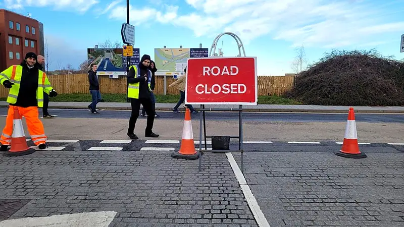 A Road Closed sign and cones across a junction, with EST staff on hand